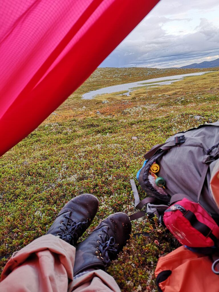 Ben och packning utanför tält med utsikt mot en sjö i fjällen.