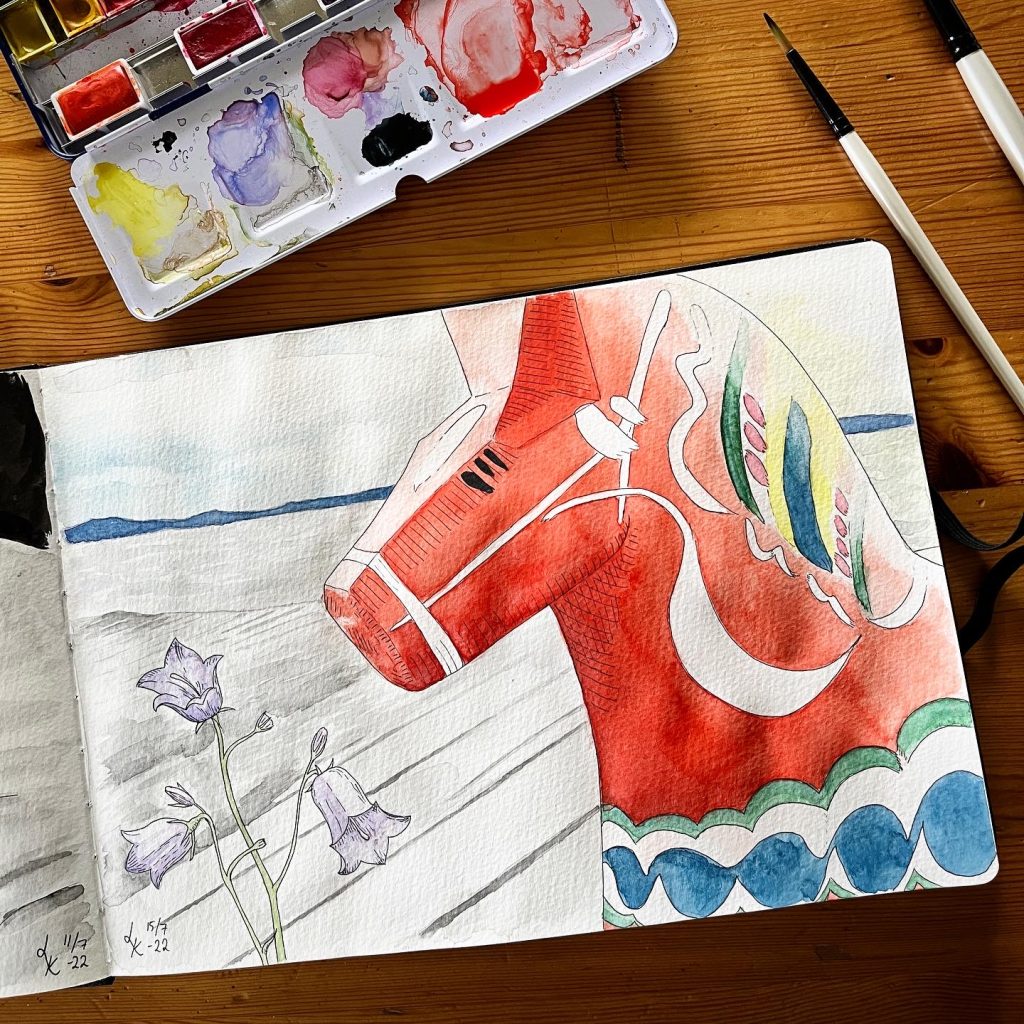Akvarellfärger och penslar bredvid sida i Lisa Krigas skissbok: dalahäst och blåklockor framför Siljan och berg i bakgrunden
